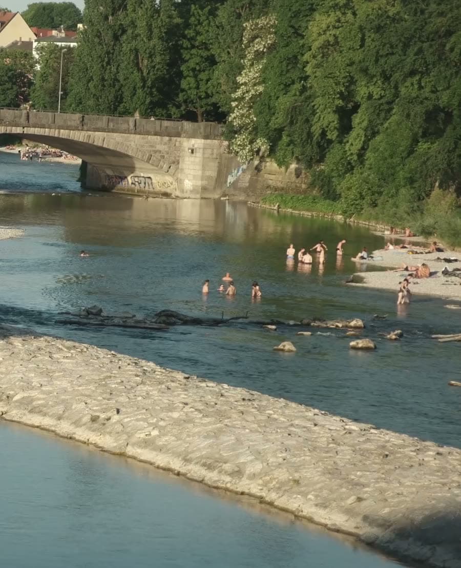 Badende in einem Seitenarm der Isar, dahinter eine steinerne Brücke