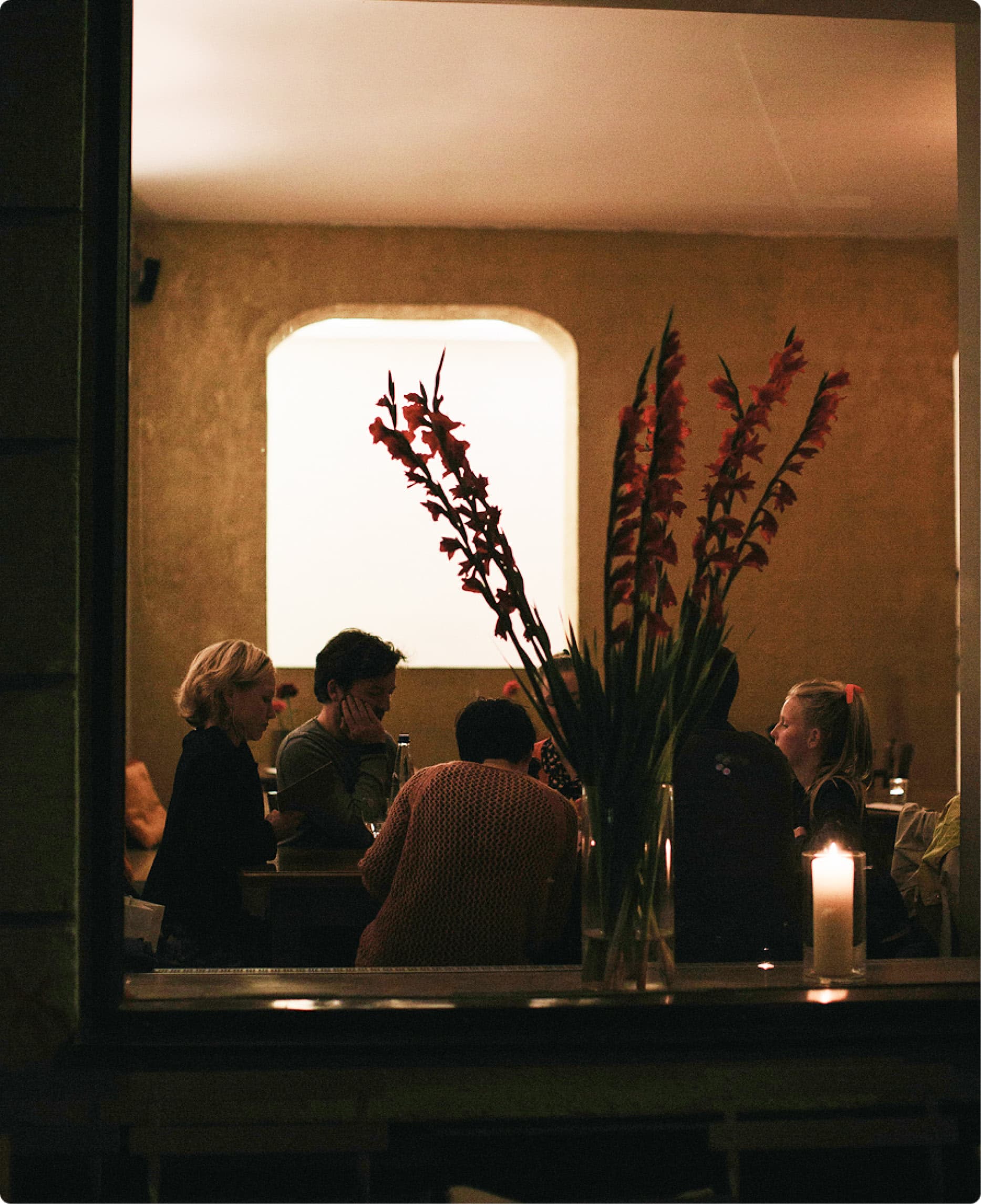 Blick durch ein Fenster in ein warm beleuchtetes Restaurant mit Gästen und Blumenschmuck