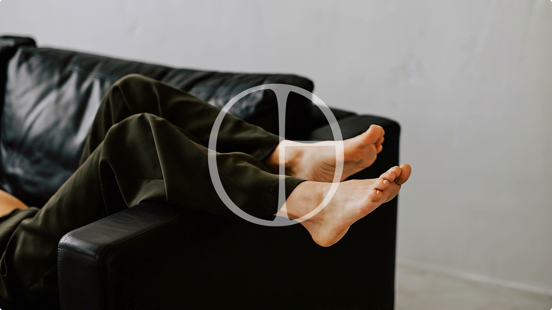 Person mit hochgelegten Beinen auf einem dunklen Ledersofa vor heller Wand