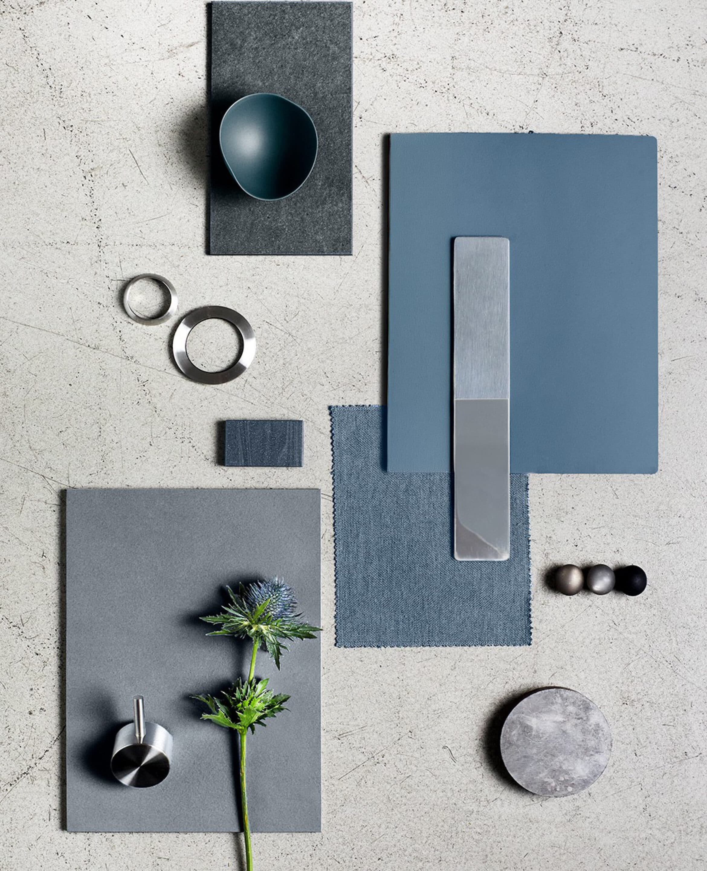 Materialcollage in Blautönen mit Steinplatten, Denimgewebe, Edelstahlringen und einer Distelblüte